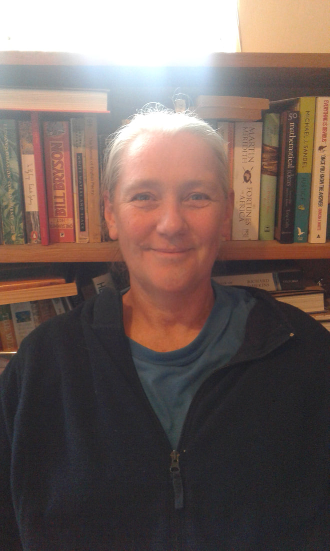 Smiling White woman with dark-blonde hair standing in front of a bookcase