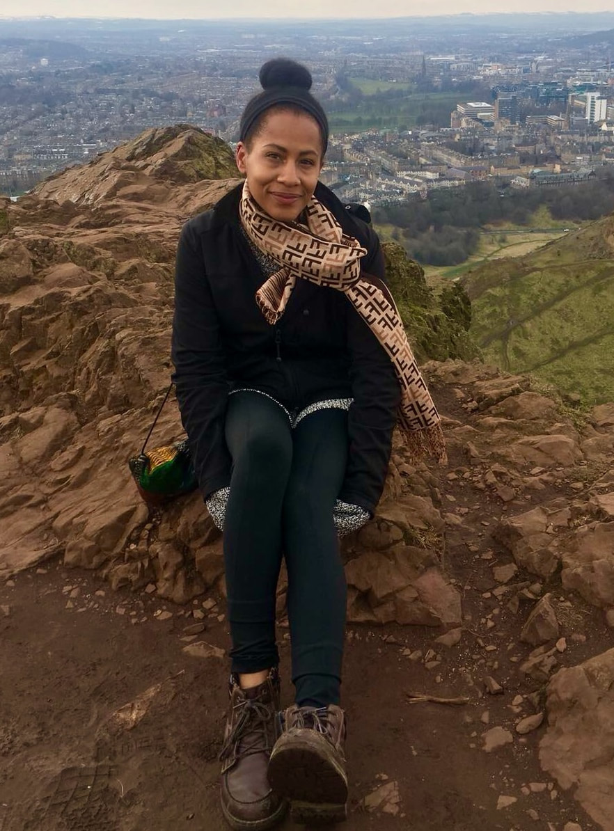 Smiling woman of color in jacket and scarf, picture taken in front of a scenic outdoor background