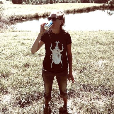 Picture shows a thin white woman with dark blond hair in a ponytail messily, jeans rolled up, converse all-stars, a black t-shirt with a large rhino beetle on it. She holds a beer can to her mouth. She is standing in a large green field with a large pond behind her.